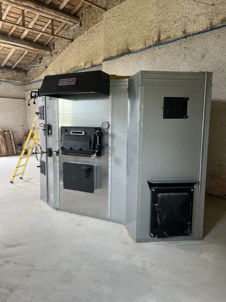 Chaudière industrielle SEBASTIA grise avec portes noires et manomètre, installée dans une salle aux murs bruts et plafond bois. Échelle XPERTY visible.