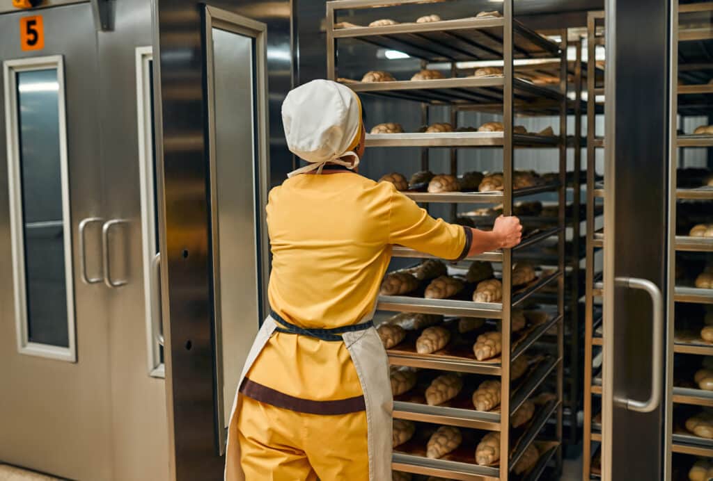 Boulanger en uniforme jaune et toque poussant un chariot de pains tressés non cuits vers un four industriel numéroté '5'.